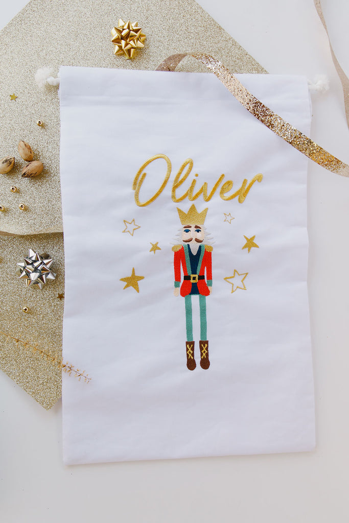 White drawstring bag with a nutcracker design and 'Oliver' text, surrounded by decorative items on a light background.